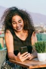 Веселая молодая кудрявая латиноамериканка болтает по мобильному телефону, отдыхая на террасе кафе в солнечный летний вечер — стоковое фото