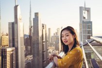 Молода азійка, мандрівниця на верхівці тераси, дивиться на камеру, яка посміхається, щоб не дивитися на місто Дубай з сучасною архітектурою. — стокове фото