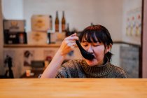 Молодая азиатка в свитере ест рамен ложкой за деревянным столиком в кафе — стоковое фото