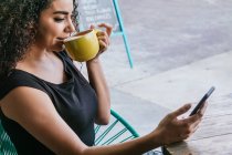 From above of happy young Hispanic female browsing mobile phone while enjoying delicious cappuccino at table of street cafe — Stock Photo