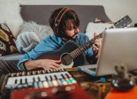 Молодой человек в наушниках играет на гитаре возле стола с ноутбуком и синтезатором дома, глядя в камеру — стоковое фото