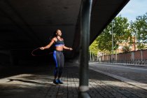 Mujer afroamericana fuerte en ropa deportiva saltando con una cuerda mientras hace ejercicio en la calle de la ciudad en un día soleado - foto de stock
