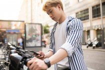Maschio etnico focalizzato utilizzando app di condivisione su smartphone e noleggio bici parcheggiato sulla strada della città — Foto stock