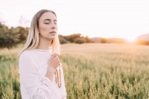 Gläubige Frau in weißem Kleid hält Perlen mit Kreuz in der Hand, während sie in der Einsamkeit auf einem ruhigen ländlichen Feld in der Natur betet — Stockfoto