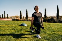 Sportsman in sportswear working out with dumbbells while performing forward lunge and breathing on meadow in city — Stock Photo