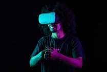 Eccitato maschio latino con acconciatura afro e occhiali VR che sperimentano la realtà virtuale su sfondo nero in studio con luci al neon — Foto stock