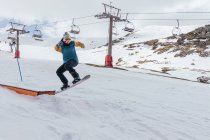 Anonymer männlicher Athlet mit Stoffmaske springt mit Snowboard über Schnee gegen Sierra Nevada und Seilbahn in Spanien — Stockfoto