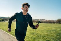 Adult male jogger in sportswear running on pavement between lawns while looking forward during training in town — Stock Photo