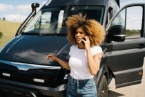 Jovem afro-americana preocupada com cabelos cacheados conversando no telefone celular enquanto pede ajuda do serviço de reparo após acidente com van campista no campo — Fotografia de Stock