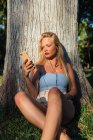 Обнаженная самка на смартфоне и слушает музыку в наушниках, сидя под деревом в парке на закате летом — стоковое фото