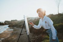Side view of young woman in stylish clothes and beret standing on grassy coast near sand and ocean in sunny day while drawing picture with brush on canvas on easel — Stock Photo