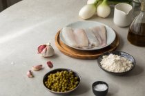 De cima dentes de alho frescos e sal colocados na mesa perto de filé de pescada, tigela com ervilhas e farinha durante a preparação de alimentos na cozinha — Fotografia de Stock