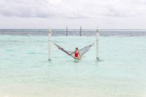 Woman in red swimsuit sitting in hammock swing over ocean surf line relaxing in Maldives on cloudy day — Stock Photo
