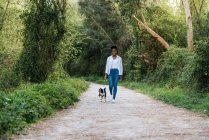 Full body of positive young African American female owner walking with loyal Border Collie dog on leash on pathway among green trees — Stock Photo