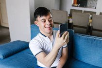 Un adolescent latino souriant atteint du syndrome de Down se faisant tirer dessus sur son smartphone alors qu'il était assis sur un canapé à la maison — Photo de stock