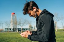 Atleta masculino barbudo en ropa deportiva y auriculares mensajería de texto en el teléfono celular mientras está de pie en el prado de la ciudad - foto de stock