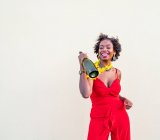 Delighted African American female in flower necklace wearing bright overall standing with bottle of champagne against white background — Stock Photo