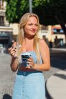 Hermosa rubia joven hembra comiendo helado frío sabroso mientras está de pie en la calle de la ciudad en el día soleado en verano - foto de stock