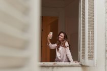 Feminino em pijama de pé perto da janela e tomando selfie celular em casa — Fotografia de Stock