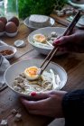 Ausgeschnittene unkenntliche Person bereitet frische gekochte Ramen-Nudeln mit Tofu, Eiern und Gemüse mit Essstäbchen auf einem Holztisch zu — Stockfoto