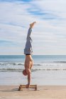 Vue latérale sur toute la longueur de l'athlète masculin exécutant handstand avec barres parallèles sur la côte sablonneuse avec des vagues de l'océan sur fond — Photo de stock