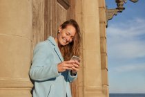 Happy adult woman in blue coat leaning on old building while browsing on cellphone in city district in sunny day under blue cloudy sky — Stock Photo