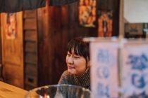 Mujer asiática en ropa casual sentada en el mostrador de madera mientras espera el orden en el bar de ramen - foto de stock