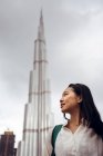 Низкий угол улыбки молодой азиатки в повседневной одежде смотрит в сторону, стоя против современной высокой башни Бурдж Халифа в Дубае в пасмурный день — стоковое фото