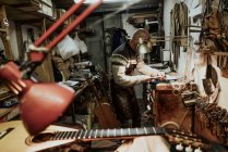 Seitenansicht eines nicht wiederzuerkennenden glatzköpfigen Kunsthandwerkers, der Instrumente an der Werkbank in einem kleinen Kunstatelier in der Nähe von akustischen Gitarren und verschiedenen Werkzeugen repariert — Stockfoto