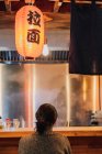 Back view of faceless black haired woman in sweater sitting at counter in cozy ramen bar — Stock Photo