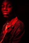 Cultivo tranquilo hombre afroamericano con estilo en chaqueta vaqueros bajo la luz roja de neón en la sombra sobre fondo negro mirando a la cámara - foto de stock