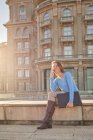 Glückliche erwachsene Frau in lässigem Outfit sitzt auf einer Steintreppe, während sie an sonnigen Tagen mit einem Ordner in der Hand in der Stadtstraße in der Nähe eines alten Gebäudes telefoniert — Stockfoto