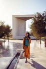 Viajera joven positiva de cuerpo completo vestida con ropa tradicional caminando por la plaza pavimentada cerca del pabellón cúbico en el Memorial de los Fundadores en Abu Dhabi mientras visita Emiratos durante las vacaciones de verano - foto de stock