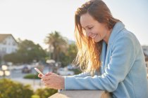 Smiling adult lady in warm coat browsing phone while leaning on fence near city street in sunny day — Stock Photo