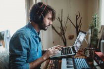 Вид сбоку молодого человека в наушниках с помощью синтезатора и ноутбука за домашним столом — стоковое фото