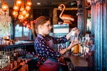 Vista lateral do barman fêmea escolhendo bebida alcoólica em garrafa para preparar coquetel no bar — Fotografia de Stock