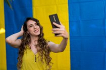 Donna con i capelli ondulati scattare selfie con il telefono cellulare mentre ride su due sfondo colorato in strada — Foto stock