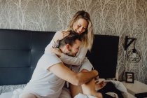 Alegre joven hombre y mujer sonriendo y abrazándose mientras están sentados en la cómoda cama en casa juntos - foto de stock