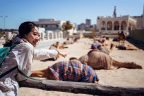 Side view of excited Asian female traveler with backpack smiling away and pointing finger on camels resting on sand place in Eastern bazaar against ancient buildings in Qatar Souq in Doha — Stock Photo
