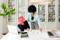 Jovens designers de moda feminina multirracial discutindo esboços para nova coleção enquanto trabalham juntos na mesa na sala de luz — Fotografia de Stock