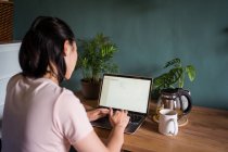 Vista trasera de la irreconocible mujer asiática freelancer leyendo documentos en el portátil mientras está sentado en la mesa durante el trabajo remoto - foto de stock