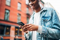 Из-под урожая веселый контент афроамериканец в джинсовой куртке серфинг современный мобильный телефон во время прогулки по городу — стоковое фото