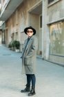 Jovem transgênero em casaco elegante e chapéu com as mãos no bolso olhando para longe à luz do dia — Fotografia de Stock