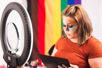 Além disso, tamanho feminino olhando para o espelho com anel de luz e aplicando maquiagem criativa contra a bandeira LGBT em estúdio — Fotografia de Stock