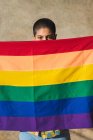 Jovem mulher étnica bissexual com cabelo curto cobrindo rosto com bandeira do arco-íris enquanto olha para a câmera no fundo bege — Fotografia de Stock