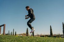 Вид сбоку спортсмена в спортивной форме, прыгающего через луг во время тренировки под голубым небом — стоковое фото