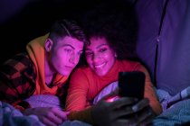 Feliz homem e mulher multirracial sorrindo descansando e navegando celular na barraca à noite — Fotografia de Stock