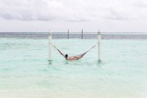 Woman in red swimsuit lying in hammock swing over ocean surf line relaxing in Maldives on cloudy day — Stock Photo