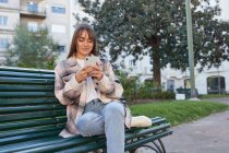 Moderna donna millenaria in elegante abito primaverile seduta su panchina e la navigazione sul telefono cellulare mentre riposava sulla strada urbana guardando altrove — Foto stock