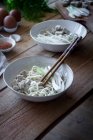 Ausgeschnittene unkenntliche Person bereitet frische gekochte Ramen-Nudeln mit Tofu, Eiern und Gemüse mit Essstäbchen auf einem Holztisch zu — Stockfoto